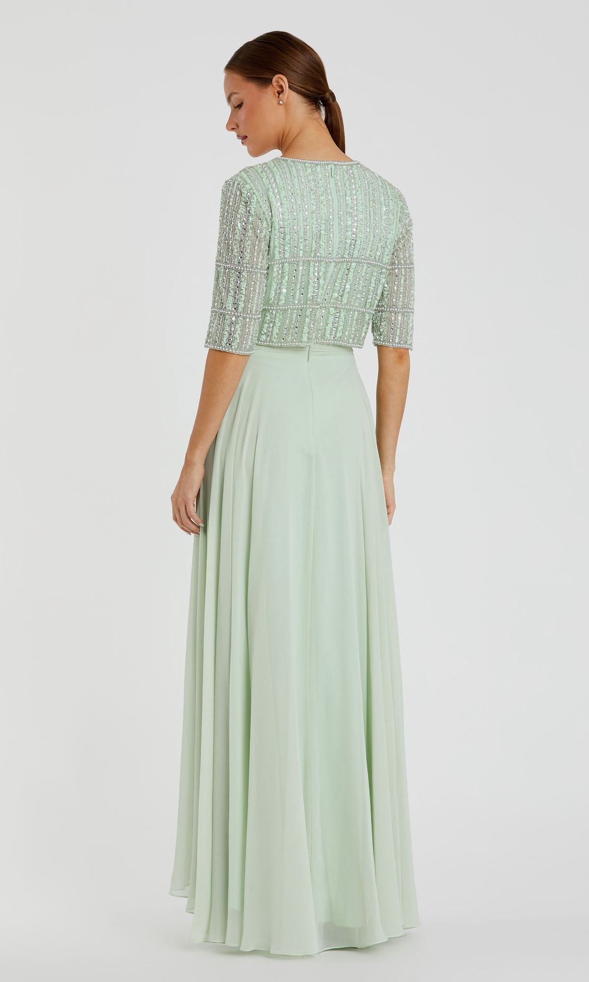 A woman with brown hair in a low ponytail stands with her back to the camera, wearing the Mac Duggal 9180—a light green, long formal dress with a beaded top and short sleeves.